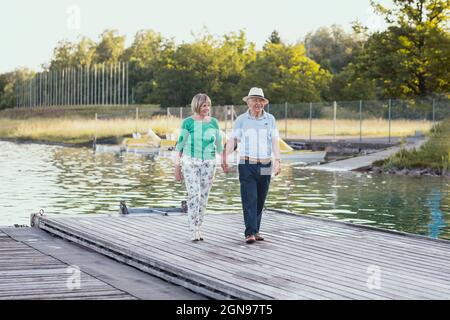 Das ältere Paar hält sich die Hände, während es auf dem Steg am See läuft Stockfoto
