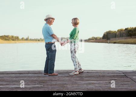 Das ältere Paar hält sich die Hände, während es auf dem Steg am See steht Stockfoto