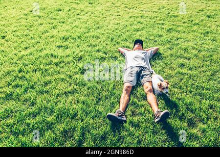 Mann entspannt sich mit den Händen hinter dem Kopf von Hund auf dem Rasen Stockfoto