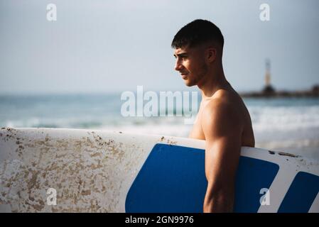 Seitenansicht eines jungen Mannes, der sein Surfbrett am Strand hält. Stockfoto