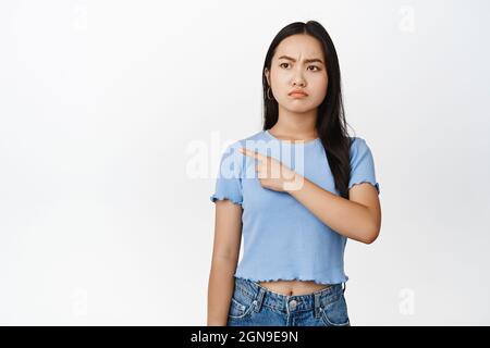 Wütendes asiatisches Mädchen, das nach links zeigt, mit mürrischem Stirnrunzeln, starren vor Abneigung und stehen im T-Shirt auf weißem Hintergrund Stockfoto