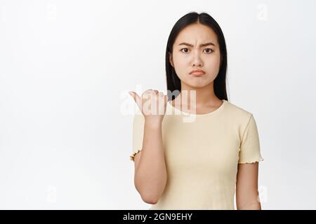 Wütendes asiatisches Mädchen sieht mürrisch oder eifersüchtig aus, Punkte auf leerem Raum, weißer Hintergrund Stockfoto