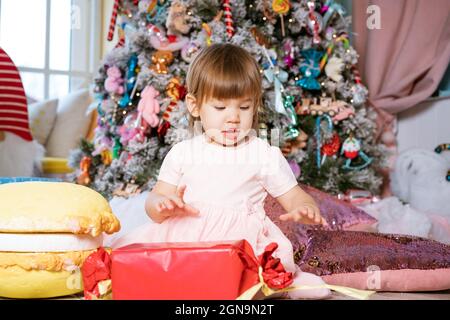 Porträt eines kleinen blonden Mädchens, das in der Nähe des Weihnachtsbaums spielt. Das Kind trägt ein weißes T-Shirt und einen rosa Rock. Urlaubskonzept Stockfoto