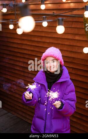 Kaukasisches Mädchen hat Spaß im Winter auf der Straße mit Wunderkerzen in der Hand lächelt vor Glück, während sie in einer warmen Fliederjacke und Strickmütze steht. Helle Funken aus den Lichtern Stockfoto