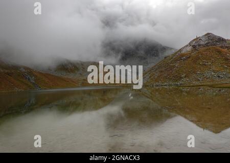 Bergsee im Herbst unter niedrigen Wolken, Vegetation in Herbstfarben, Berge spiegeln sich in spiegelglattem Wasser Stockfoto