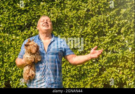 Reifer Mann mit Yorkshire Terrier Hund unter Spritzwasser an heißen Tag im Garten. Stockfoto