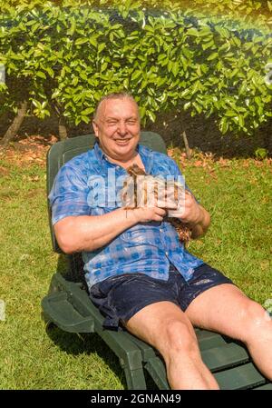 Reifer Mann mit Yorkshire Terrier Hund unter Spritzwasser an heißen Tag im Garten. Stockfoto