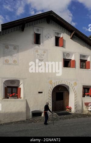 Ein älterer Bewohner spaziert mit einem Stock an der Vorderseite eines der alten Häuser in Scuol Sot vorbei, der Altstadt, die in der Nähe des alpinen Inn oder in Scuol, Graubünden oder Graubünden, Schweiz, erbaut wurde. Das restaurierte Gehöft trägt zwei Daten: 1724 und 1986, das Jahr, in dem es renoviert wurde. Die Fassade ist mit Sgraffito-Dekorationen und einer Inschrift in Rätoromanisch geschmückt, einer Vermächtnis-Sprache des Römischen Reiches, die noch immer von etwa der Hälfte der Bewohner dieses Kurortes im Unterengadiner Tal gesprochen wird. Stockfoto