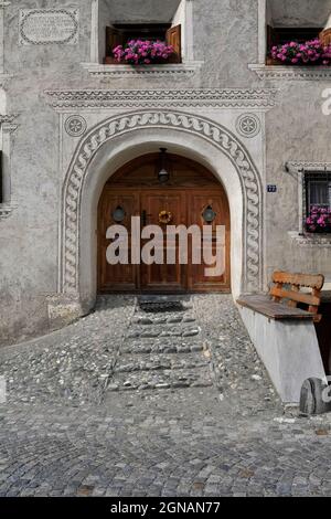 Ein gestufte gepflasterter Weg führt zur Eingangstür des Hauses 72 in der Altstadt von Scuol in Graubünden oder Kanton Graubünden, Schweiz. Die breite, runde Veranda des restaurierten Familienhauses ist von wirbelnden dekorativen Sgraffito begrenzt und der Weg wird von einer geschnitzten Holzbank flankiert, die an einem Sockel befestigt ist. Auf der Vorderseite befindet sich auch eine Inschrift in romanisch, einer alten Sprache, die noch von vielen Einheimischen gesprochen wird, und ein Fries mit schuppigen Meereslebewesen und einer Meerjungfrau. Stockfoto