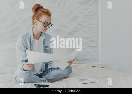 Junge konzentrierte Frau Hausfrau in Brille Verwaltung Familienbudget, während auf dem Bett zu Hause sitzen Stockfoto