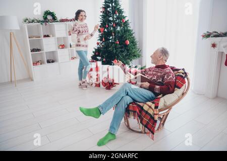 Foto von charmanten niedlichen Alter Paar gekleidet drucken Pullover genießen Buch Dekoration Baum lächelnd drinnen Zimmer Haus Stockfoto