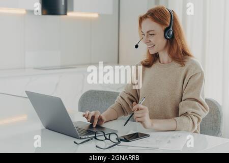 Happy Redhead Woman sieht sich das Online-Webinar an und verwendet ein Headset, das auf den Laptop-Bildschirm fokussiert ist Stockfoto