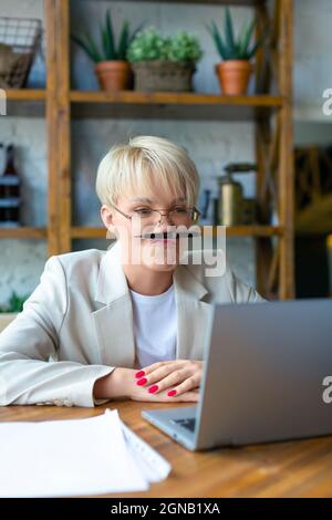 Foto einer jungen Blondine mit Brille vor einem Laptop. Sie ist es leid, während eines Online-Geschäftsvideoanrufs zu arbeiten und herumzutäuschen. Arbeiten mit Stockfoto