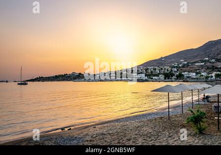 Landschaftlich schöner Sonnenuntergang mit dem Strand im Vordergrund und dem Dorf im Hintergrund. Reflexionen auf dem Wasser. Foinikas Syrou, Kykladen, Griechenland. Stockfoto