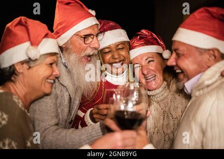 Verschiedene ältere Menschen, die am Heiligabend mit Wein jubeln und Weihnachtsmützen tragen - weicher Fokus auf afrikanisches Frauengesicht Stockfoto