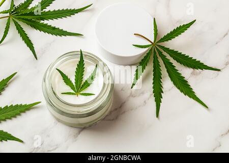 Glas mit Hautpflege-Feuchtigkeitscreme und frischen grünen Cannabis-Sativa-Blättern auf einem Marmorschreibtisch. Kräuterhanf Kosmetik Konzept. Medizinische Marihuanapflanze. Stockfoto