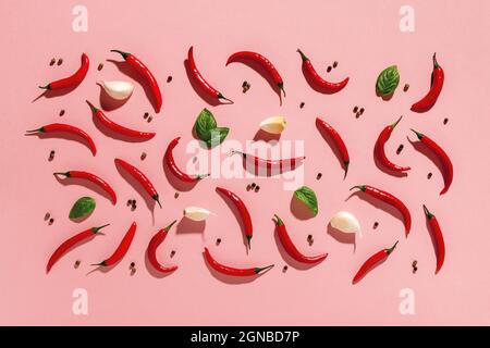 Rohe rote Chili-Paprika, wenige Knoblauchzehen und frische grüne Basilikumblätter, schwarze Pfefferkörner werfen harte Schatten auf einen pastellfarbenen korallenrosa Hintergrund. Stockfoto