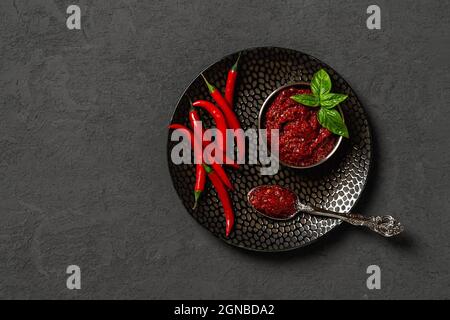 Dicke scharfe Chilisauce in einer runden Metallschüssel und eine Menge frischer roter Chilischoten auf einer schwarzen strukturierten Platte auf dunkelgrauem Hintergrund. Gewürze. Stockfoto