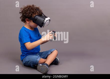 Der 4-5-jährige kaukasische Junge sitzt auf dem Boden mit einer Virtual-Reality-Brille und Game-Controllern in den Händen Stockfoto