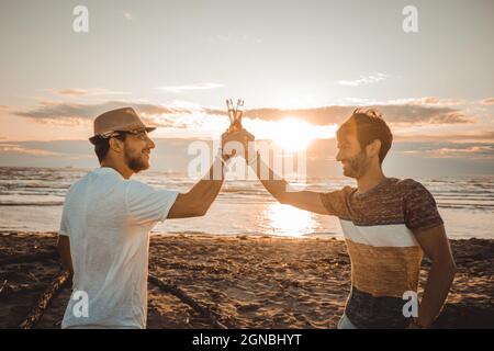 Fröhliche junge Leute verbringen Zeit zusammen am Strand und trinken Bier und toasten am Strand im Urlaub in der Dämmerung Sommer Sonnenuntergang Stockfoto