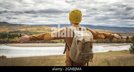 Der Mann in einem gelben Hut und Pullover mit Rucksack steht mit den nach den Seiten ausgestreckten Armen vor dem Fluss, dem herbstlichen goldenen Wald und Stockfoto