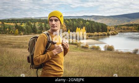 Der Mann, der in einem gelben Hut und Pullover mit Rucksack unterwegs ist, steht vor dem Fluss, dem herbstlichen goldenen Wald und den Hügeln. Konzept der Freiheit Stockfoto