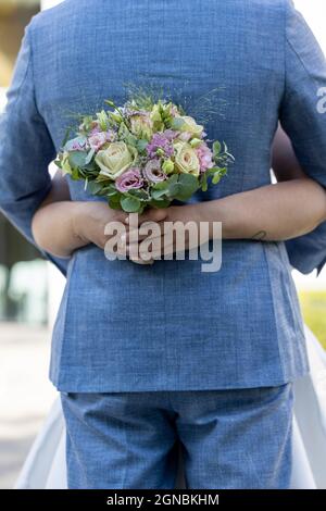 Nahaufnahme der Hand einer Braut mit einem Bouquet aus weißen und rosa Rosen, die die blaue Weste des Bräutigams auf einem Wiesenhintergrund umarmen. Hochwertige Fotos Stockfoto