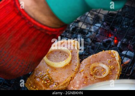 Zwei Schweinesteaks mit Zwiebelringen auf dem Grill und brennende Kohlen mit einer Hand in einem roten Handschuh im Vordergrund. Hochwertige Fotos Stockfoto