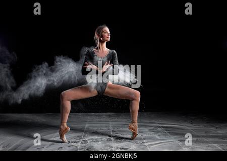 Flexible Frau tanzt mit Mehl auf schwarzem Hintergrund im Studio, bewegt sich mit Beinen auseinander. Talentierter Athlet Balletttänzer im Bodysuit in Zeitlupe, lo Stockfoto