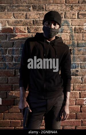 afroamerikanischer Mann in Maske, der an einer Ziegelwand steht, Hooligan in schwarzer Kleidung. Schwarzer Kerl stellen eine Gefahr für die Gesellschaft dar, warten auf Opfer, begehen ein Verbrechen. Stockfoto