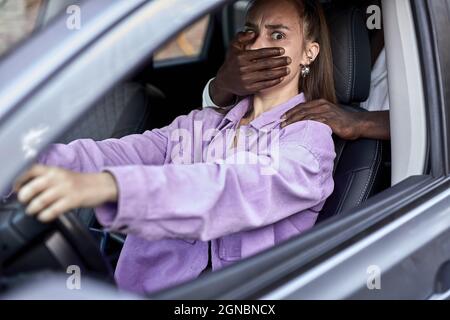 Gefährlicher schwarzer Mann, der verängstigte junge Frau im Auto bedroht, nicht erkennbarer afrikanischer Mann schließt den Mund einer verängstigten Frau und raubt. S Stockfoto