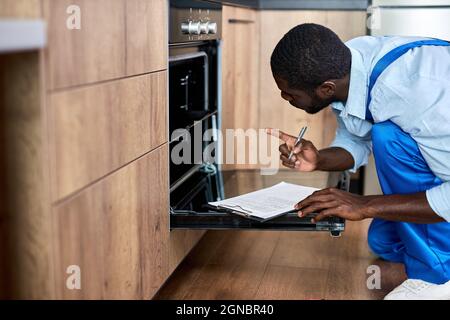 Nice Contractor Handyman in Overalls Blick in den elektrischen Ofen Untersuchung vor der Reparatur, während Schreiben von Informationen über Ofen, Zeigen Index fing Stockfoto