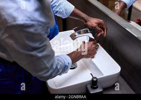 Nahaufnahme von afrikanischer schwarzer Klempnerhand, die versucht, einen Wasserhahn im Badezimmer zu öffnen, repariert ein nicht erkennbarer Handwerker oder Klempner in blauer Uniform Insta Stockfoto