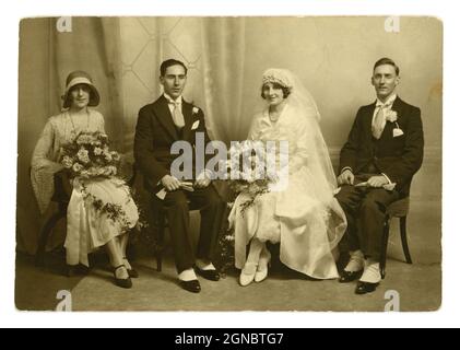 Originaler Vintage-Fotoklassiker aus den 1920er Jahren, Flapper-Ära, Hochzeitsgruppenportrait, attraktives verheiratetes Paar, Paare - schöne Braut, Bräute mit langem Schleier, weißes Brautkleid, atemberaubender Kopfschmuck, neben dem Bräutigam sitzend und bester Mann, der passende Anzüge mit geflügelten Hemdkragen und Spatzen trägt. Hübscher Blumenstrauß, Blumensträuße. Die Brautjungfer trägt einen modischen Cloche-Hut. Mann und Frau aus den 1920er Jahren. Ehemänner und Ehefrauen. Circa 1924, Großbritannien Stockfoto
