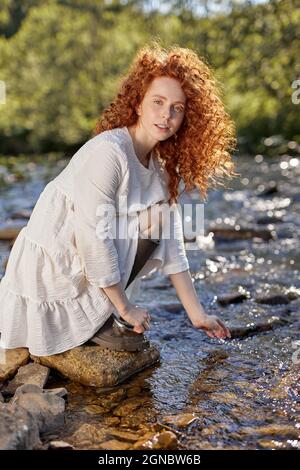 Schöne junge rothaarige lockige Frau sitzt neben dem Fluss berühren Wasser, in der Natur am Sommertag, trägt weißes Kleid, charmante verträumte Dame allein. Por Stockfoto