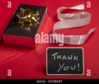 Komposition auf einem roten Hintergrund eines Geschenks mit Kakade, Band und Tafel mit den Worten „Danke!“ Stockfoto