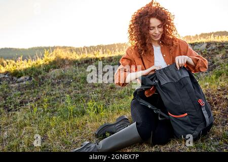 Die rothaarige lockige Frau sitzt auf einem Rucksack, der sich auf Gras öffnet und ruht sich während einer Wanderung auf den Bergen aus, und macht eine Pause. Porträt einer jungen schönen kaukasischen Dame in Stockfoto