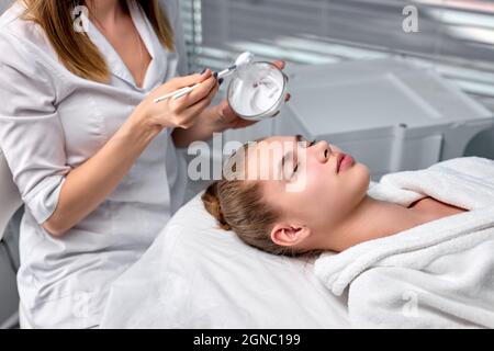 Beschnittene Kosmetologin, die Maske aus der Schüssel auf das hübsche Frauengesicht aufsetzt, wunderschöne kaukasische Frau im Spa, die Gesichtsbehandlungen hat, mit geschlossenen Augen liegend Stockfoto