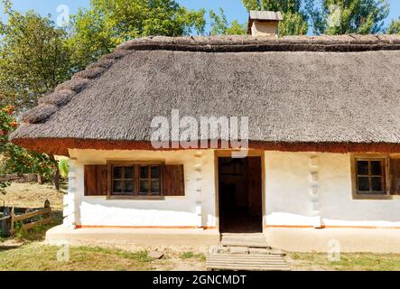 Fragment der Fassade eines alten traditionellen ukrainischen Landhauses mit Reetdach vor dem Hintergrund eines Sommergartens und blauen Himmels. Stockfoto