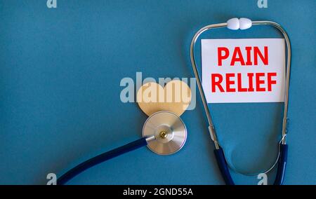 Symbol für Schmerzlinderung. Weiße Karte mit Worten Schmerzlinderung, schöner blauer Hintergrund, Holzherz und Stethoskop. Medizin und Schmerzlinderung Konzept. Stockfoto