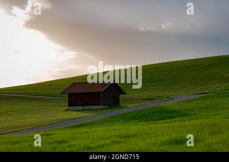 Dies ist eine Landschaftsaufnahme einer kleinen bayerischen Hütte auf einem Hügel in der Nähe einer kleinen Straße. Es ist Sommer im Allgäu. Stockfoto