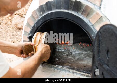 Handwerklich hergestellter Holzofen und Männerhände, die mit einem Blasebalg in die Glut blasen Stockfoto