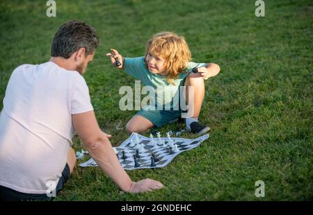 Glückliche Familie von Vater und Sohn Junge spielen Schach auf grünem Gras im Park im Freien, Schachspiel Stockfoto