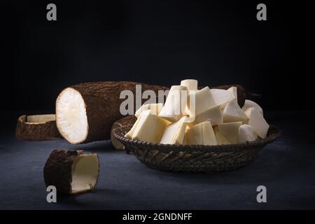 Cassava oder Manihot, auch bekannt als Manioc, Yuca oder Brazillian Pfeilwurzel, Wurzelgemüse in Stücke geschnitten bereit zum Kochen mit einer halbgeschnittenen Wurzel, auf dunkel Stockfoto