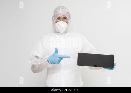 Ein Mann trägt Schutzkleidung mit Gesichtsmaske, Handschuhen und Schutzanzug, der eine Schachtel hält Stockfoto