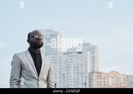 Horizontale Porträtaufnahme eines selbstbewussten, hübschen schwarzen Mannes, der einen eleganten grauen Anzug und eine Brille trägt, die im Freien steht und wegschaut Stockfoto