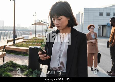 Horizontale, mittlere Porträtaufnahme einer modernen asiatischen Frau, die im Freien steht und auf dem Heimweg nach Abschluss der Arbeit eine SMS an jemand schreibt, der sein Smartphone verwendet Stockfoto