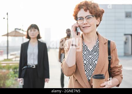 Horizontale, mittlere Porträtaufnahme einer schönen, reifen Frau mit lockigen roten Haaren, die nach dem Arbeitstag mit jemandem auf dem Heimweg telefoniert Stockfoto