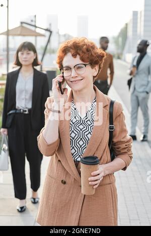 Vertikale mittellange Porträtaufnahme einer fröhlichen, reifen Frau mit lockigen roten Haaren, die auf der Straße unterwegs ist und mit jemandem telefoniert, nachdem sie den Arbeitstag beendet hat Stockfoto