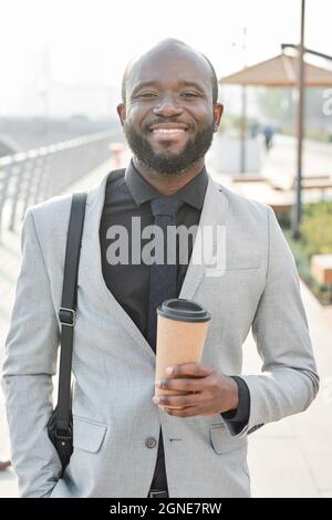 Vertikales mittleres Porträt eines fröhlich schönen afroamerikanischen Mannes, der nach Beendigung der Arbeit im Büro eine Kaffeetasse im Freien hält Stockfoto